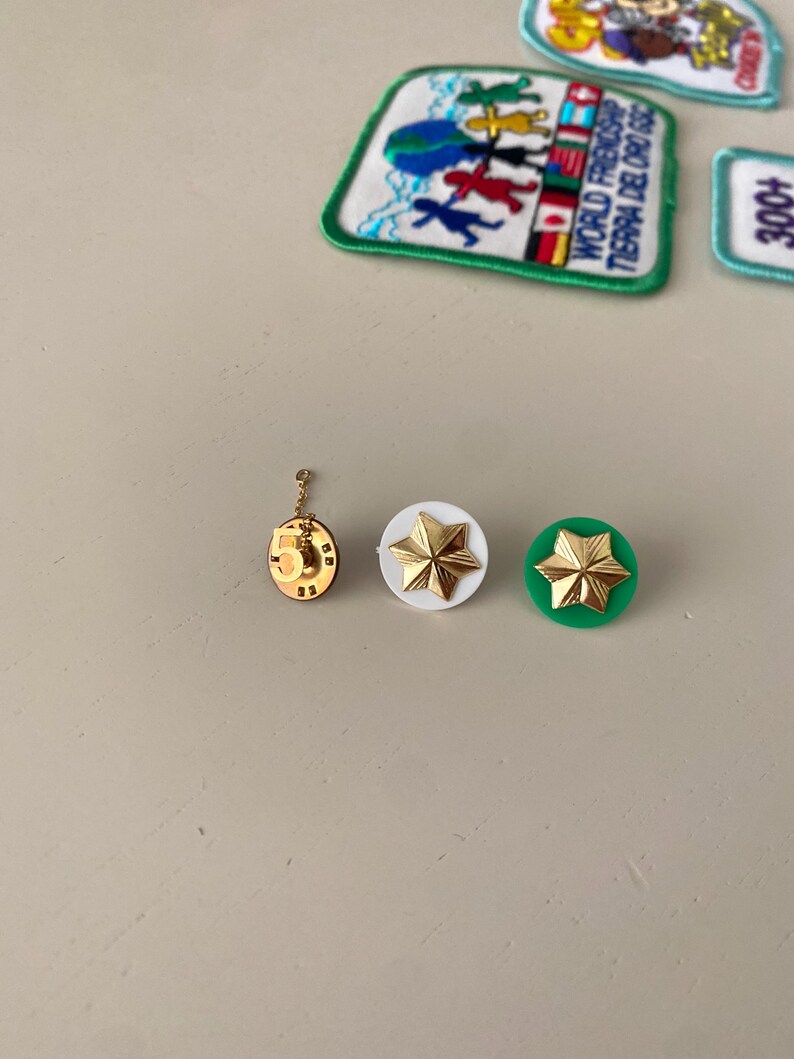 May include: Three gold-colored pins with a star design. The star pins are on a white and green background. The pin on the left has a number "5" on it. The pin in the middle is on a white background. The pin on the right is on a green background.