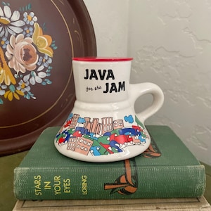 Puede incluir: Taza de cerámica blanca con tapa roja y un diseño de escena de ciudad colorida. La taza tiene el texto "JAVA for the JAM" impreso en la parte delantera.