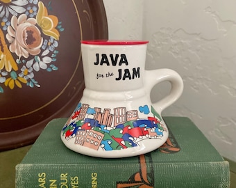 May include: White ceramic mug with a red lid and a colourful city scene design. The mug has the text "JAVA for the JAM" printed on the front.