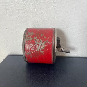 May include: A vintage red Twin Plex pencil sharpener with a silver crank and trim. The sharpener has the words "Twin Plex" in a white, stylized font. The red paint shows signs of wear, giving it a rustic appearance.