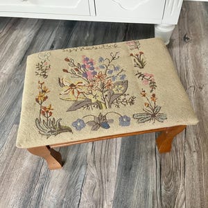 May include: A small, rectangular footstool with a beige fabric top featuring a floral needlepoint design in shades of pink, blue, yellow, and green. The wooden legs are a warm brown colour. The footstool is a decorative home decor item.