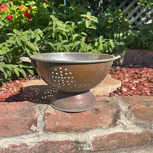 May include: A round metal colander with a weathered copper-toned finish, featuring a wide pedestal base, two side handles, and a series of small circular drainage holes arranged in a grid pattern across the bowl and the base of the kitchen tool.