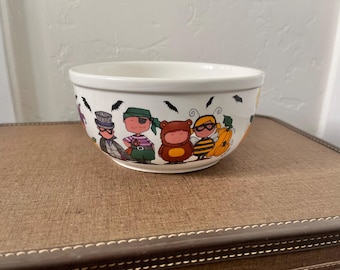 May include: White ceramic bowl with a Halloween-themed design. The bowl features cartoon characters in costumes, including a pirate, a bear, and a bee. Black bats are also included in the design. The bowl is sitting on a brown surface.