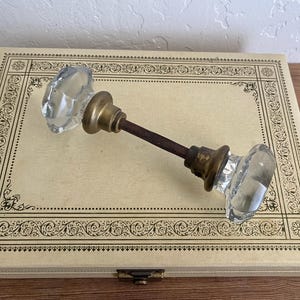 May include: A clear, faceted glass door knob set featuring two octagonal glass handles connected by a central threaded metal spindle. Each glass knob is mounted on a circular brass base, with the metal spindle extending between the two components.