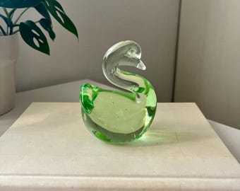 May include: A clear and green glass swan figurine. The swan has a curved neck and a rounded body, with a light green hue in the body. The figurine is sitting on a book with the words "CARPE DIEM" on the cover.