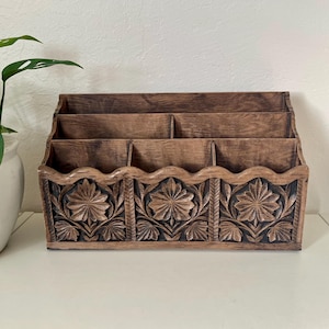 Peut inclure: Un organiseur de bureau en bois avec une finition marron foncé. L'organisateur comporte trois compartiments et un motif floral sculpté décoratif en bas. Une plante en pot est visible en arrière-plan.