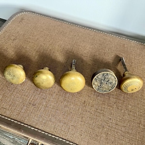 May include: Five antique brass door knobs and pulls in various designs. The knobs are in shades of gold and cream, with a variety of shapes, including round and cylindrical. The knobs are arranged on a brown surface.