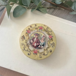 May include: A round, pale yellow metal tin featuring a central circular illustration of two figures in historical attire. The lid is decorated with gold-toned scrollwork and pink rose accents surrounding the central image of the couple.