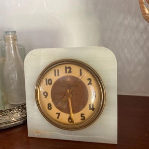 May include: A vintage white marble clock with a brass face and black hands. The clock face has Roman numerals and the words "Westclox" and "Made in USA".
