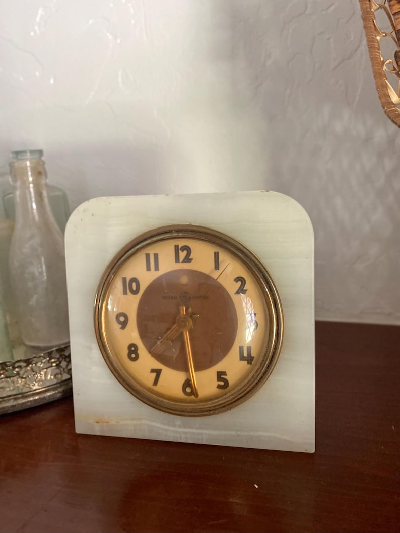May include: A vintage white marble clock with a brass face and black hands. The clock face has Roman numerals and the words "Westclox" and "Made in USA".