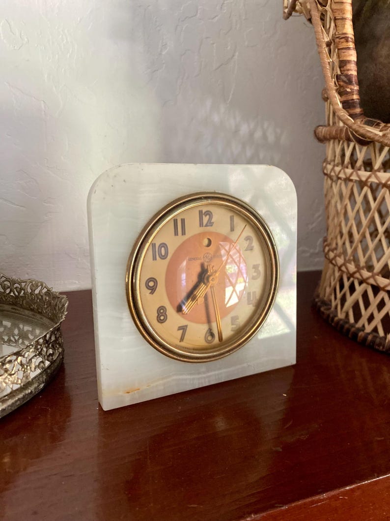 May include: A white marble clock with a gold-toned face and black hands. The clock has Roman numeral markings and a small, round, orange-colored center.