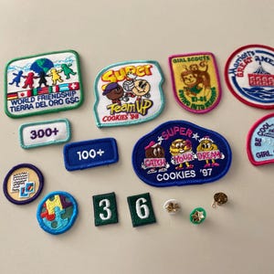 May include: A collection of Girl Scout cookie badges featuring various designs and colors. The badges include "Super Cookies '97", "World Friendship", "Be Cool, Be a Reader", "Girl Scouts '96-'97", and "Swim Into Reading".