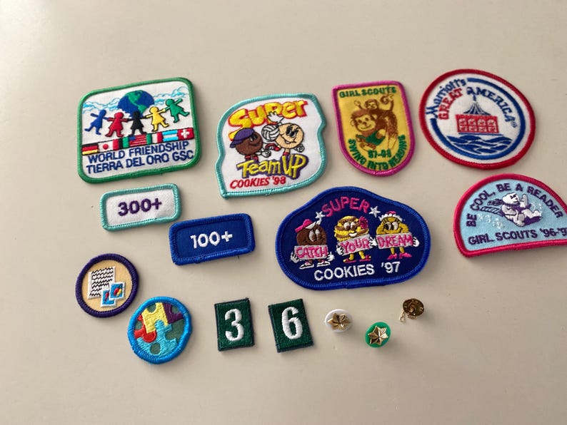 May include: A collection of Girl Scout cookie badges featuring various designs and colors. The badges include "Super Cookies '97", "World Friendship", "Be Cool, Be a Reader", "Girl Scouts '96-'97", and "Swim Into Reading".