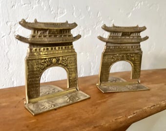 May include: A pair of gold-toned metal bookends shaped like traditional Chinese pagodas. The bookends are detailed with intricate carvings and are sitting on a wooden surface.