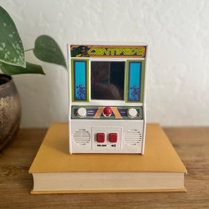 Può includere: Un cabinato arcade Centipede bianco con schermo, pulsanti e il logo del gioco. Il gioco è appoggiato su un libro giallo, con una pianta sullo sfondo. Il gioco ha un design retrò con accenti verdi, blu e rossi.