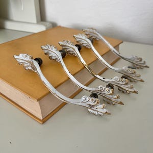 Könnte beinhalten: Sechs antike Schrankgriffe in Weiß und Gold mit aufwendigen, blattartigen Verzierungen. Die Griffe haben ein gebogenes Design und sind mit schwarzen Schrauben befestigt. Die Beschläge sind gealtert und weisen Abnutzungserscheinungen auf.