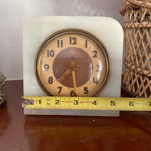 May include: A vintage white marble clock with gold accents and a round face. The clock face has black Roman numeral hour markers and black hands. The clock is 1 inch tall.