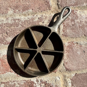 May include: A round cast iron pan with a textured dark brown surface, featuring six triangular compartments arranged in a radial pattern and a single handle with a teardrop-shaped opening at the end for hanging or storage purposes.