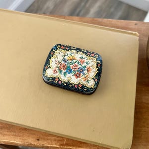 Op de afbeelding: Een klein, vierkant, vintage blikje met een bloemenpatroon. Het blikje is zwart met gouden accenten en heeft een kleurrijk bloemenpatroon met rode, gele, blauwe en groene bloemen.
