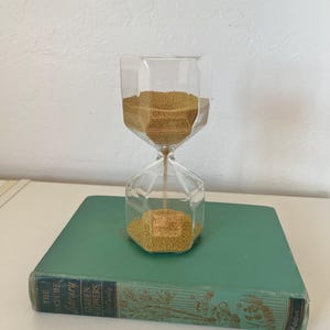 May include: A clear, geometric hourglass filled with golden sand, resting on a vintage teal book. The hourglass has a faceted design, and the sand is concentrated in the top and bottom chambers. The book's cover features gold lettering and floral illustrations.
