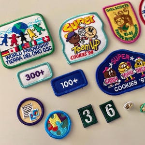 May include: A collection of colorful embroidered patches featuring various themes, including Girl Scouts, cookies, and world friendship. The patches are arranged on a white surface with some numbers and a few small pins.