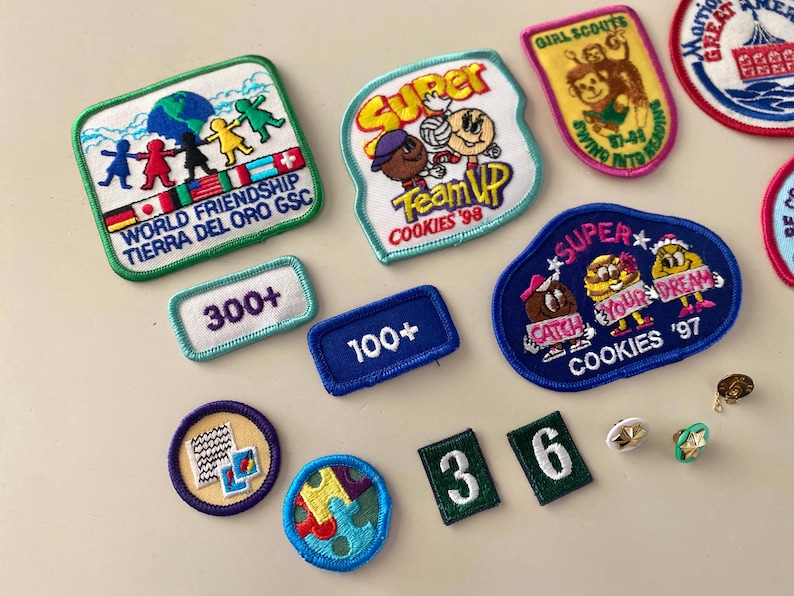 May include: A collection of colorful embroidered patches featuring various themes, including Girl Scouts, cookies, and world friendship. The patches are arranged on a white surface with some numbers and a few small pins.