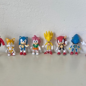 Könnte beinhalten: Eine Sammlung von acht farbenfrohen Sonic the Hedgehog-Charakterfiguren. Die Figuren umfassen Sonic, Tails, Amy Rose, Super Sonic, Mighty, Metal Sonic, Knuckles und Dr. Eggman. Jede Figur ist etwa 7.6 cm groß und steht auf einer weißen Oberfläche.