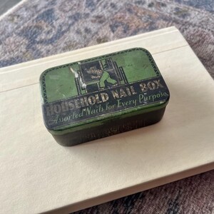 May include: Vintage rectangular metal nail box with a green and black design. The box reads "HOUSEHOLD NAIL BOX Assorted Nails for Every Purpose". The box is resting on a cream-colored book.