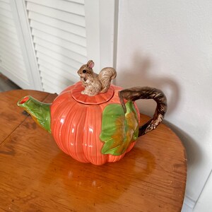 May include: A ceramic teapot shaped like a pumpkin, with an orange body and green accents. A small squirrel figurine sits atop the lid. The handle is brown and the spout is green. A decorative item for the kitchen or dining room.