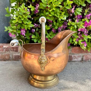 Puede incluir: Antiguo brasero de carbón de cobre con base y asa de latón. El brasero presenta un detalle de cabeza de león y asas de cerámica. El cobre tiene una pátina cálida y envejecida. La pieza está sobre un fondo de vegetación.