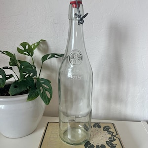 Pode incluir: Uma garrafa de vidro transparente com fecho basculante, com uma rolha branca com vedação de borracha vermelha. A garrafa tem um gargalo estreito e um corpo arredondado. O ano "1895" está gravado na garrafa. A garrafa está em cima de um livro.