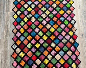 Puede incluir: Una colorida manta de ganchillo hecha a mano. La manta presenta un patrón repetitivo de cuadrados multicolores, incluyendo rojo, amarillo, verde y rosa, sobre un fondo negro. Los bordes también son negros, creando un borde definido.