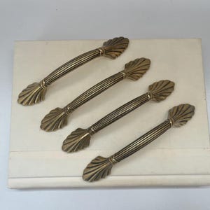 May include: Four antique brass cabinet pulls with a shell design on the ends. The pulls have a ribbed center and are arranged on a cream-colored surface. These decorative pulls are for furniture restoration.