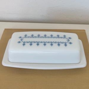 May include: A white rectangular butter dish with a matching lid. The lid features a blue floral design. The dish is placed on a white rectangular base, and sits on a light brown surface.