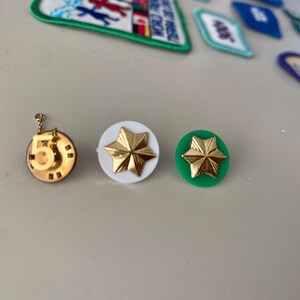 May include: Three gold-colored pins with different designs. The first pin is a circle with the number 5 in the center. The second pin is a white circle with a gold star in the center. The third pin is a green circle with a gold star in the center.
