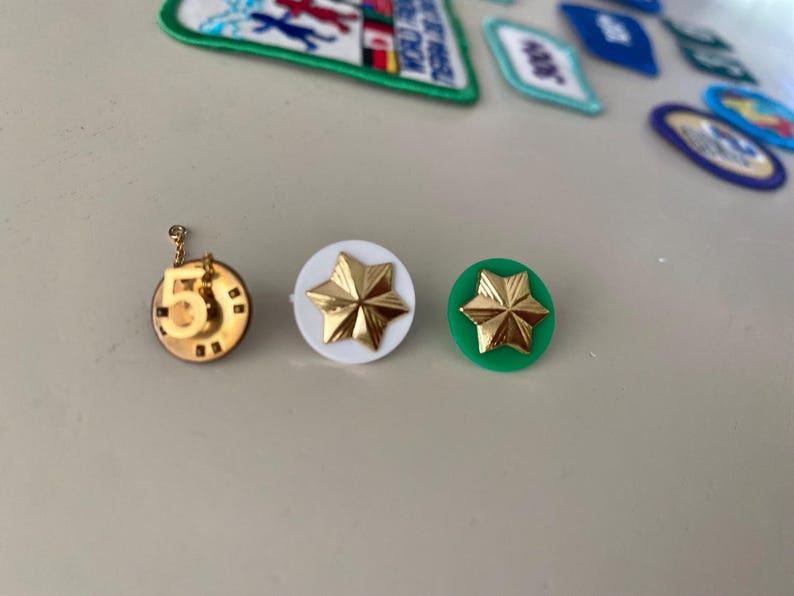 May include: Three gold-colored pins with different designs. The first pin is a circle with the number 5 in the center. The second pin is a white circle with a gold star in the center. The third pin is a green circle with a gold star in the center.