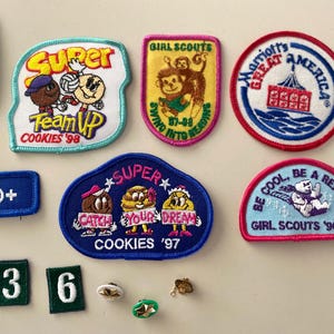May include: A collection of Girl Scout cookie badges featuring various designs and slogans, including "Super Team Up Cookies '98", "Super Catch Your Dream Cookies '97", "Girl Scouts 57-88 Swimming Into Reading", "Marriott's Great America", "Be Cool, Be A Reader Girl Scouts '96-'97", and "Friendship Girl Scouts".