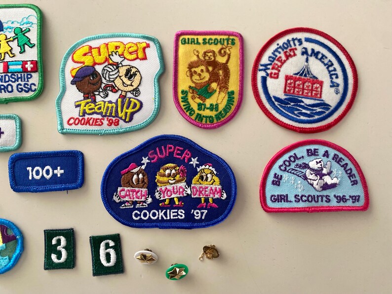 May include: A collection of Girl Scout cookie badges featuring various designs and slogans, including "Super Team Up Cookies '98", "Super Catch Your Dream Cookies '97", "Girl Scouts 57-88 Swimming Into Reading", "Marriott's Great America", "Be Cool, Be A Reader Girl Scouts '96-'97", and "Friendship Girl Scouts".
