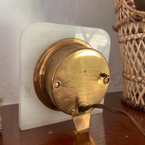 May include: A vintage General Electric clock with a brass face and a white alabaster base. The clock has a black cord and a label that reads "General Electric Clock, Model 203-A, Volts 115, 60 Cycles, Made in USA, General Electric Co., Bridgeport, Conn., U.S.A."