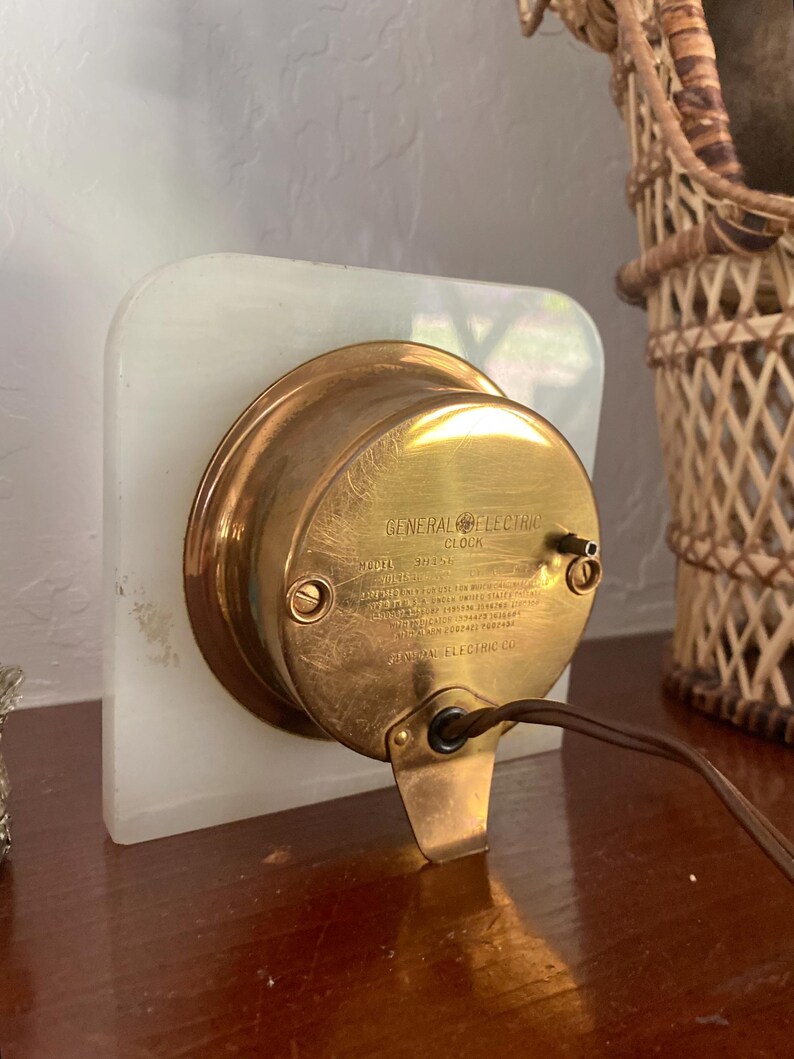 May include: A vintage General Electric clock with a brass face and a white alabaster base. The clock has a black cord and a label that reads "General Electric Clock, Model 203-A, Volts 115, 60 Cycles, Made in USA, General Electric Co., Bridgeport, Conn., U.S.A."