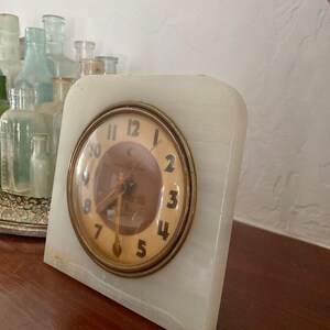 May include: A white marble clock with gold-toned hands and Roman numeral markings. The clock face is surrounded by a gold-toned ring.