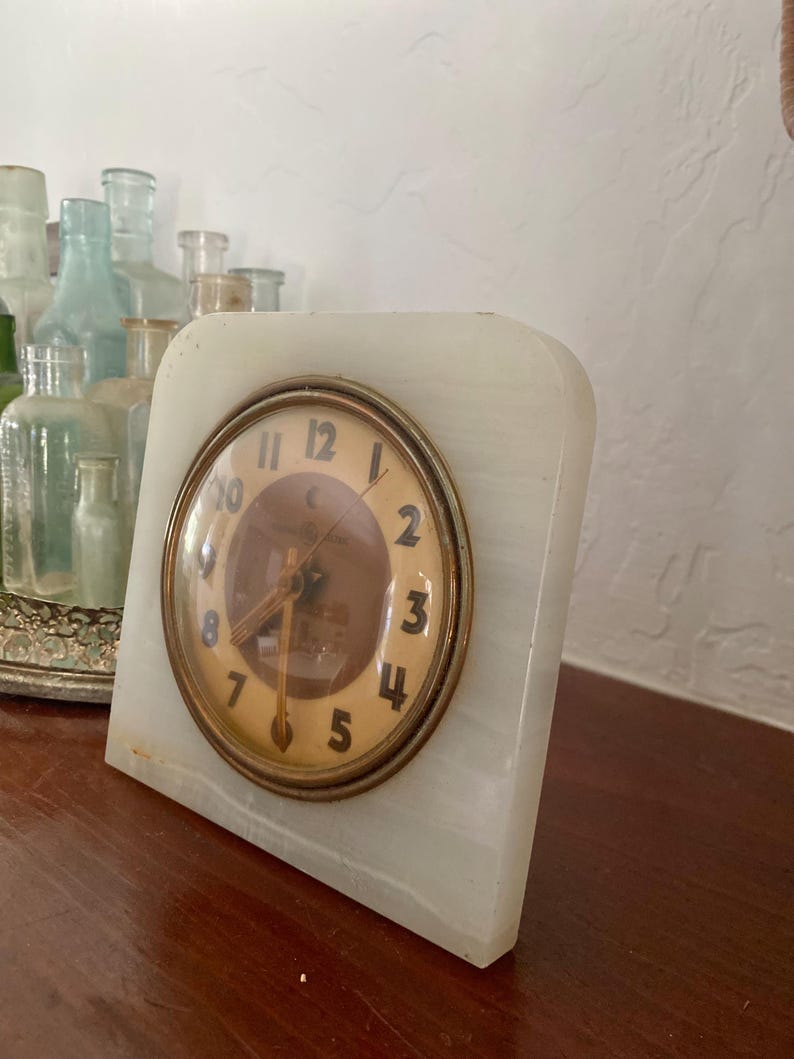 May include: A white marble clock with gold-toned hands and Roman numeral markings. The clock face is surrounded by a gold-toned ring.