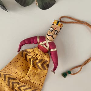 May include: Handmade Indian puppet doll with a painted face, wearing a yellow patterned skirt and a red top. The doll has a gold crown and gold accents. A brown string is attached to the doll's head.