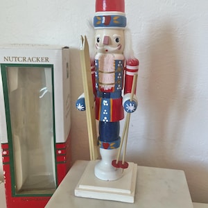 Könnte beinhalten: Handbemalte Nussknacker-Figur aus Holz im Ski-Thema. Der Nussknacker trägt einen roten Hut, eine blaue und rote Jacke und eine blaue Hose. Er hält Skier und Skistöcke. Eine Schachtel mit der Aufschrift "Nussknacker" befindet sich im Hintergrund.