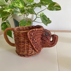 Puede incluir: Una cesta de mimbre marrón con forma de elefante con asa. La cesta está hecha de ratán tejido y tiene un acabado natural.