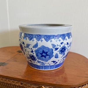 Op de afbeelding: Een ronde, blauw-witte keramische bloempot met een bloemenmotief. De pot heeft een brede rand en een licht uitlopende vorm. Het blauwe bloemenpatroon is geplaatst tegen een witte achtergrond. De pot staat op een houten oppervlak.