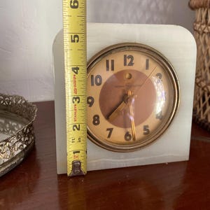 May include: A white alabaster clock with a gold-toned face and black hands. The clock face has the words "General Electric" and "Life Guard" printed on it. The clock is 4 inches tall.