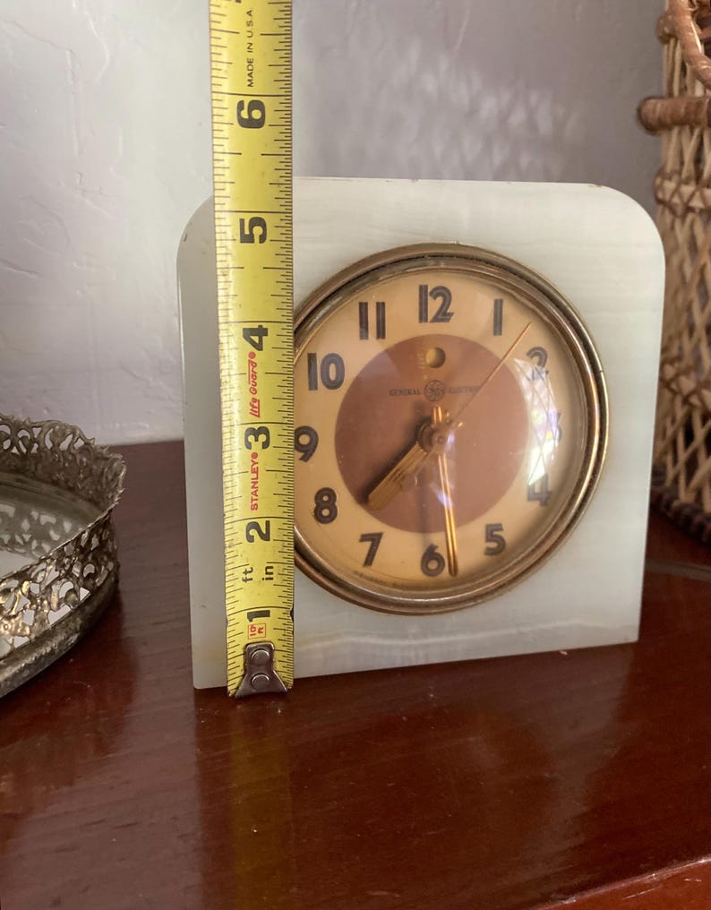 May include: A white alabaster clock with a gold-toned face and black hands. The clock face has the words "General Electric" and "Life Guard" printed on it. The clock is 4 inches tall.