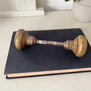 Peut inclure: Ensemble de boutons de porte anciens en laiton avec une tige de connexion, posé sur un livre bleu foncé. Les boutons ont une forme arrondie et une patine chaude et vieillie. Une pièce de quincaillerie vintage.