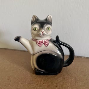 May include: A ceramic teapot shaped like a cat, with a black and white body, green eyes, and a pink bow tie. The teapot has a handle and a spout, and is sitting on a brown surface.